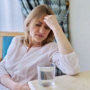 Headache, migraine, depression of mature woman sitting at home on chair with glass of water. Stress, health problems, illness, age people concept