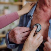 Doctor visiting senior man and examinig him with stethoscope indoors at home, listening to heartbeat.