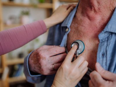 Doctor visiting senior man and examinig him with stethoscope indoors at home, listening to heartbeat.