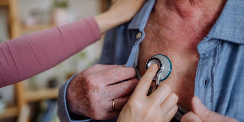 Doctor visiting senior man and examinig him with stethoscope indoors at home, listening to heartbeat.