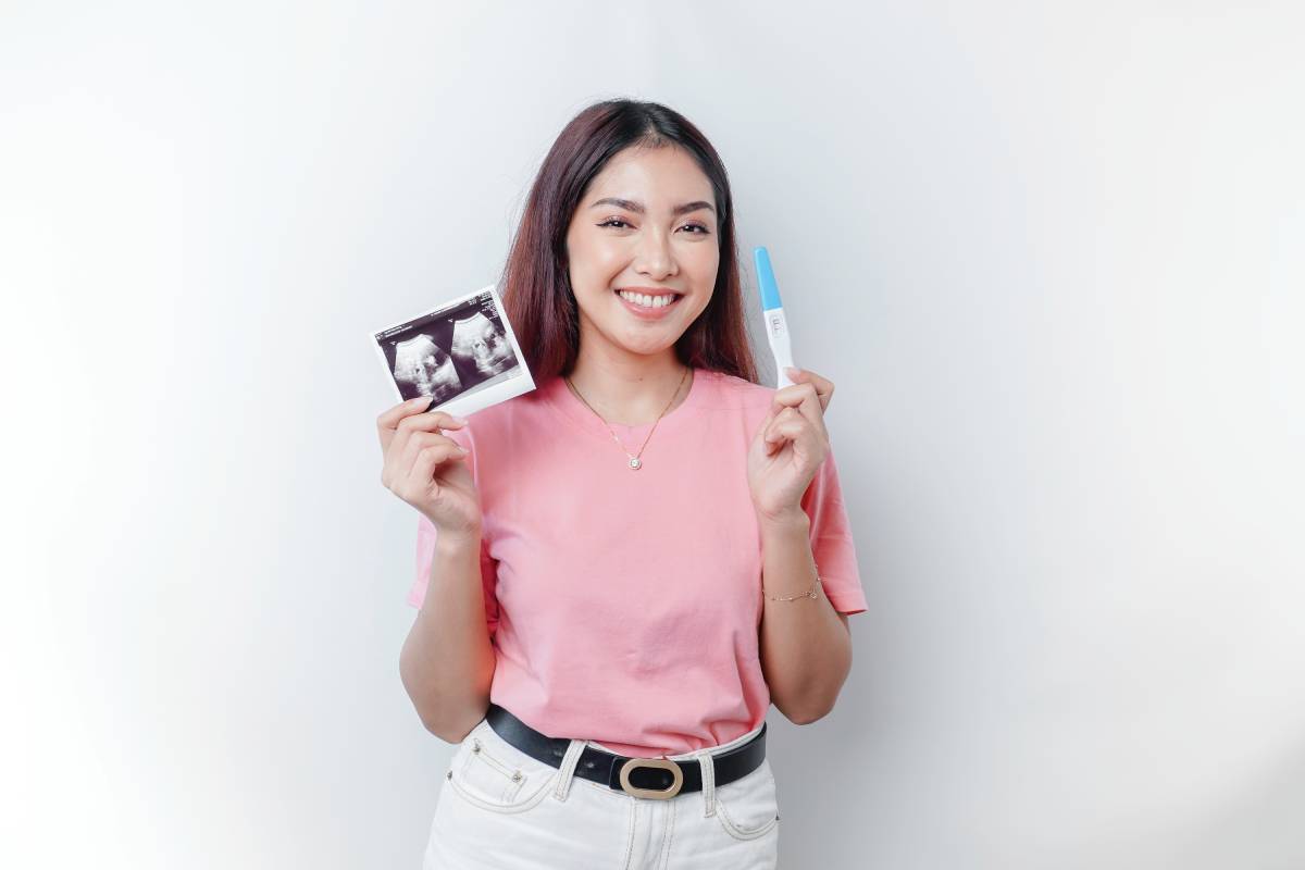 Happy young woman wearing pink t-shirt showing her pregnancy test and ultrasound picture.