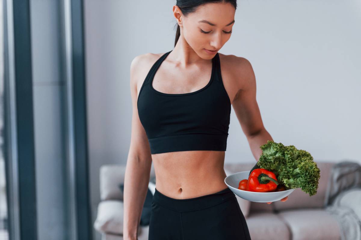 holds healthy food in hands. Young woman with slim body shape in black sportswear have fitness day indoors at home