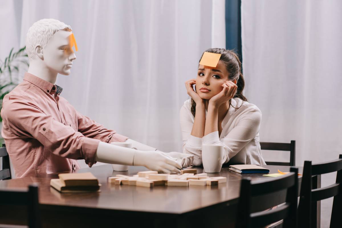 sad young woman with sticky note on forehead sitting at table with mannequin, loneliness concept
