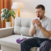 Unhappy middle aged man suffering from fever and flu, blowing nose in napkin, sitting in living room interior. Covid-19 lockdown, treatment of illness, cold and runny
