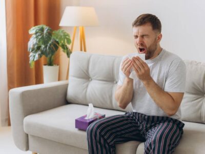 Unhappy middle aged man suffering from fever and flu, blowing nose in napkin, sitting in living room interior. Covid-19 lockdown, treatment of illness, cold and runny