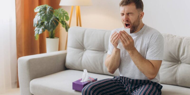 Unhappy middle aged man suffering from fever and flu, blowing nose in napkin, sitting in living room interior. Covid-19 lockdown, treatment of illness, cold and runny